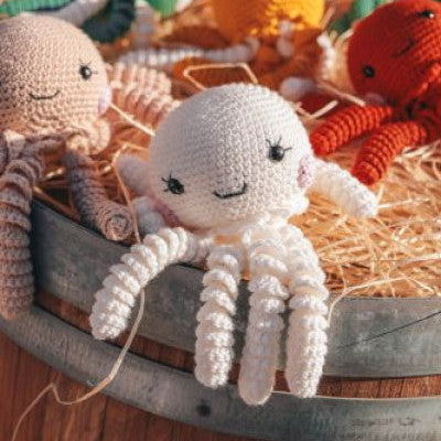 Plusieurs Doudous Manaho O'méa en crochet, dont un blanc et un beige, posés sur de la paille, mettant en avant leur texture douce et leurs tentacules.